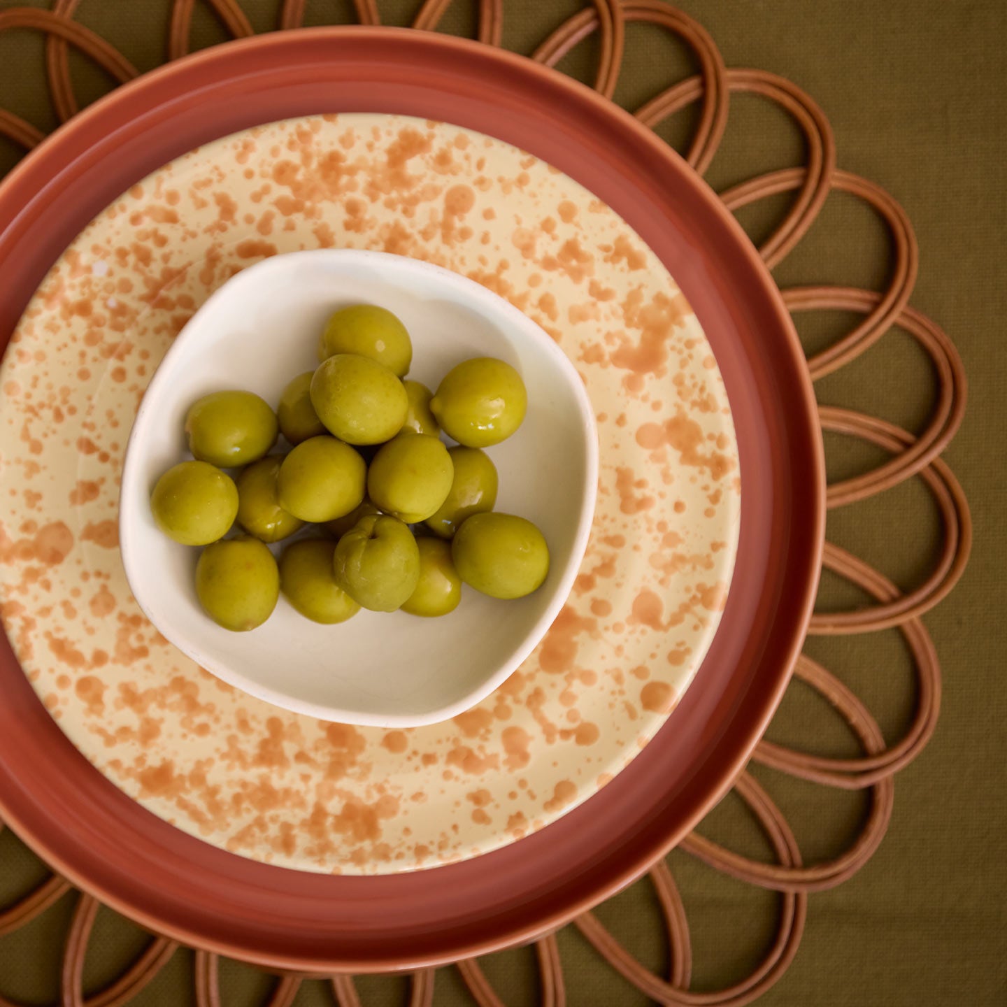 Overhead of olives in small bowl on splatter salad plate, essential dinner plate, and rattan placemat.
