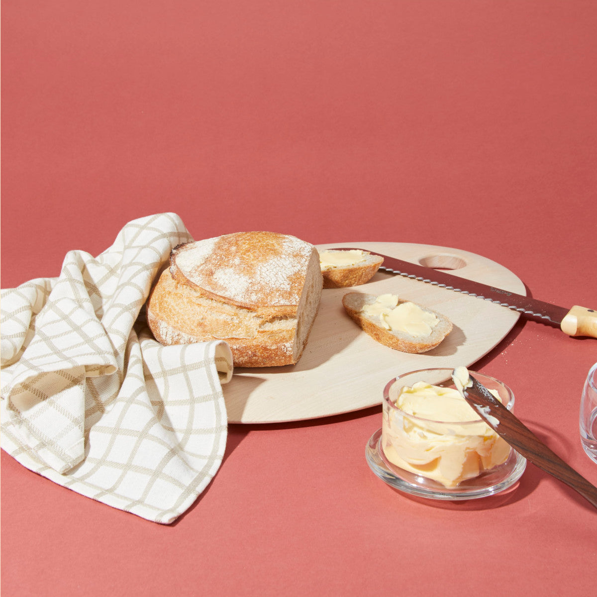 A loaf of bread, a bread knife, slices of bread with butter, a butter dish, and a cloth napkin on a round wooden cutting board against a red background.