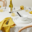 Dining table setting with white plates, mustard napkins, and glasses of white wine.