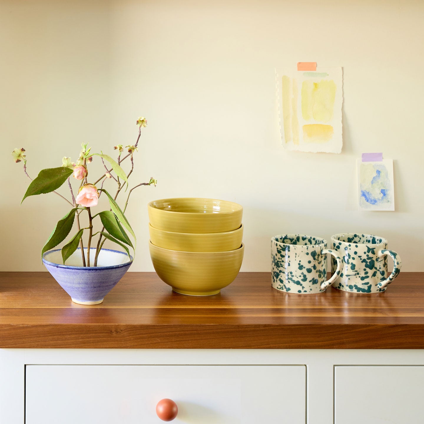 Countertop with vase and flowers, splatterware mugs, cereal bowls in mustard, and watercolor paintings on the wall.