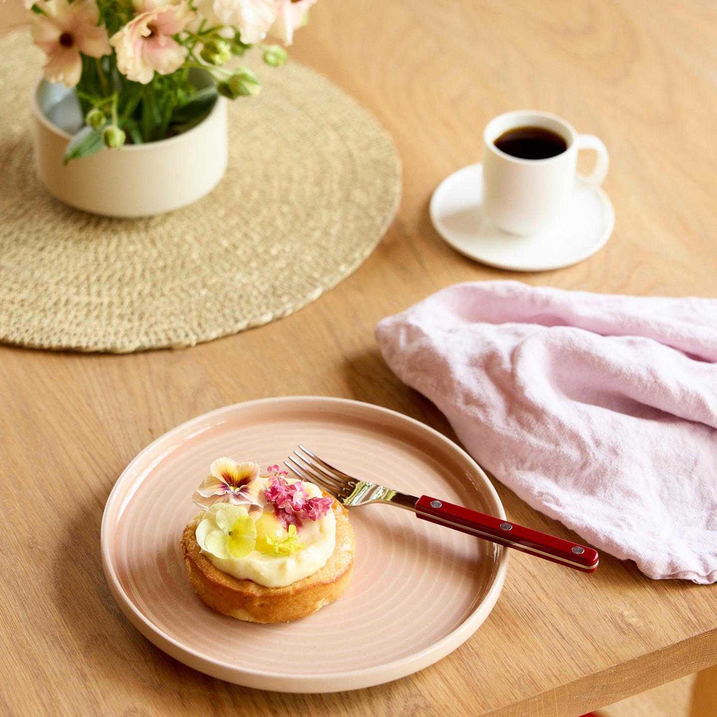 Small dessert on a pink plate with a fork, coffee cup, and flowers on a wooden table.