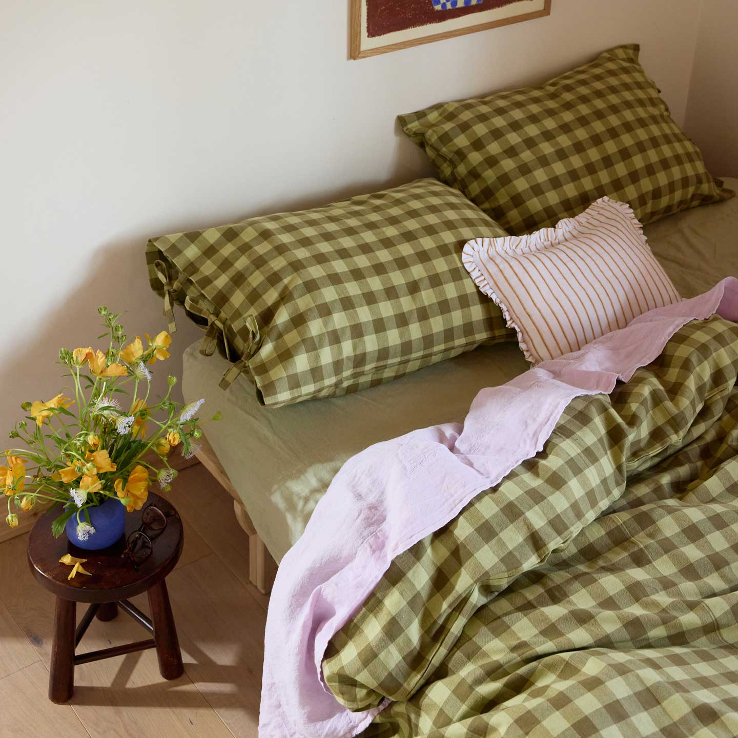 Bed with olive gingham bedding and pillows, next to a small wooden table with flowers.