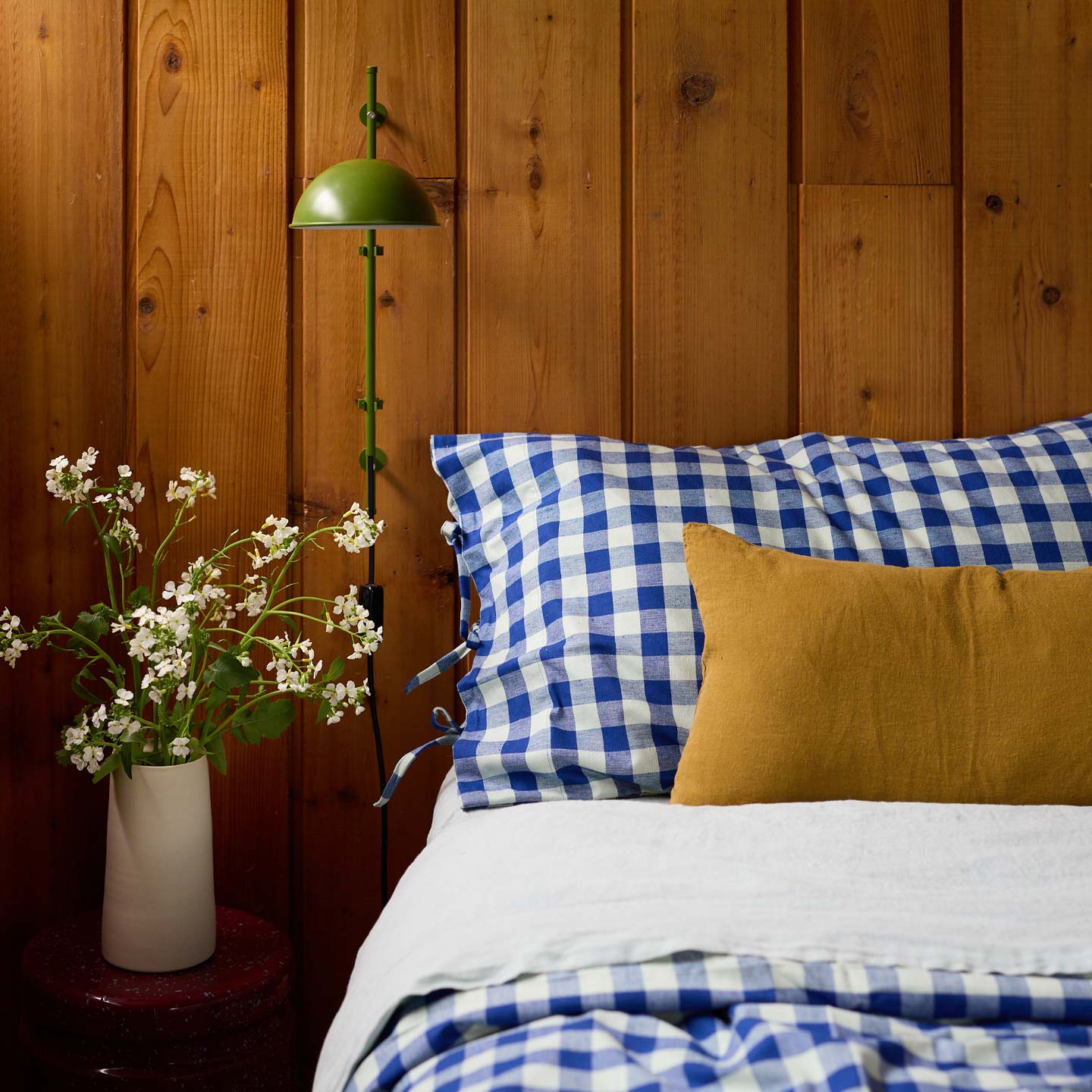 Bed with Essential Gingham Bedding in blue and white checkered pillows and a mustard yellow pillow against a wooden wall with a green lamp.