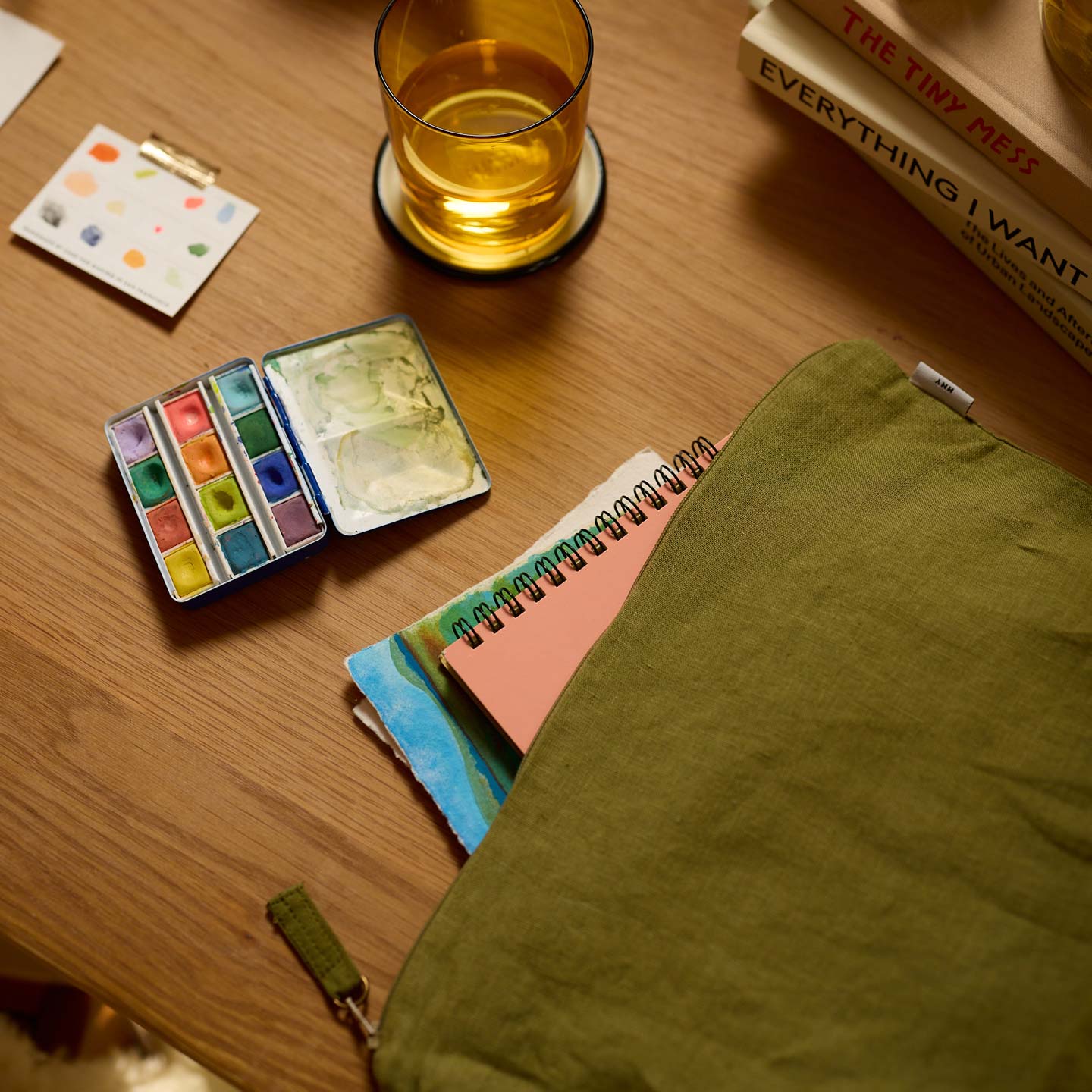 Art supplies including watercolor paints, a glass of beer, and books on a wooden table.