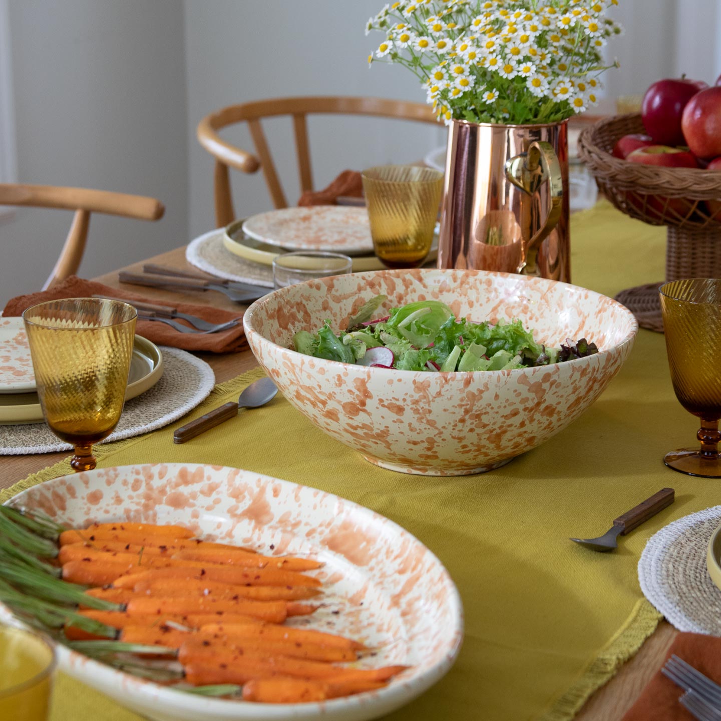 Table with mustard table runner, place settings, salad bowl, platter with carrots, and pitcher with flowers.