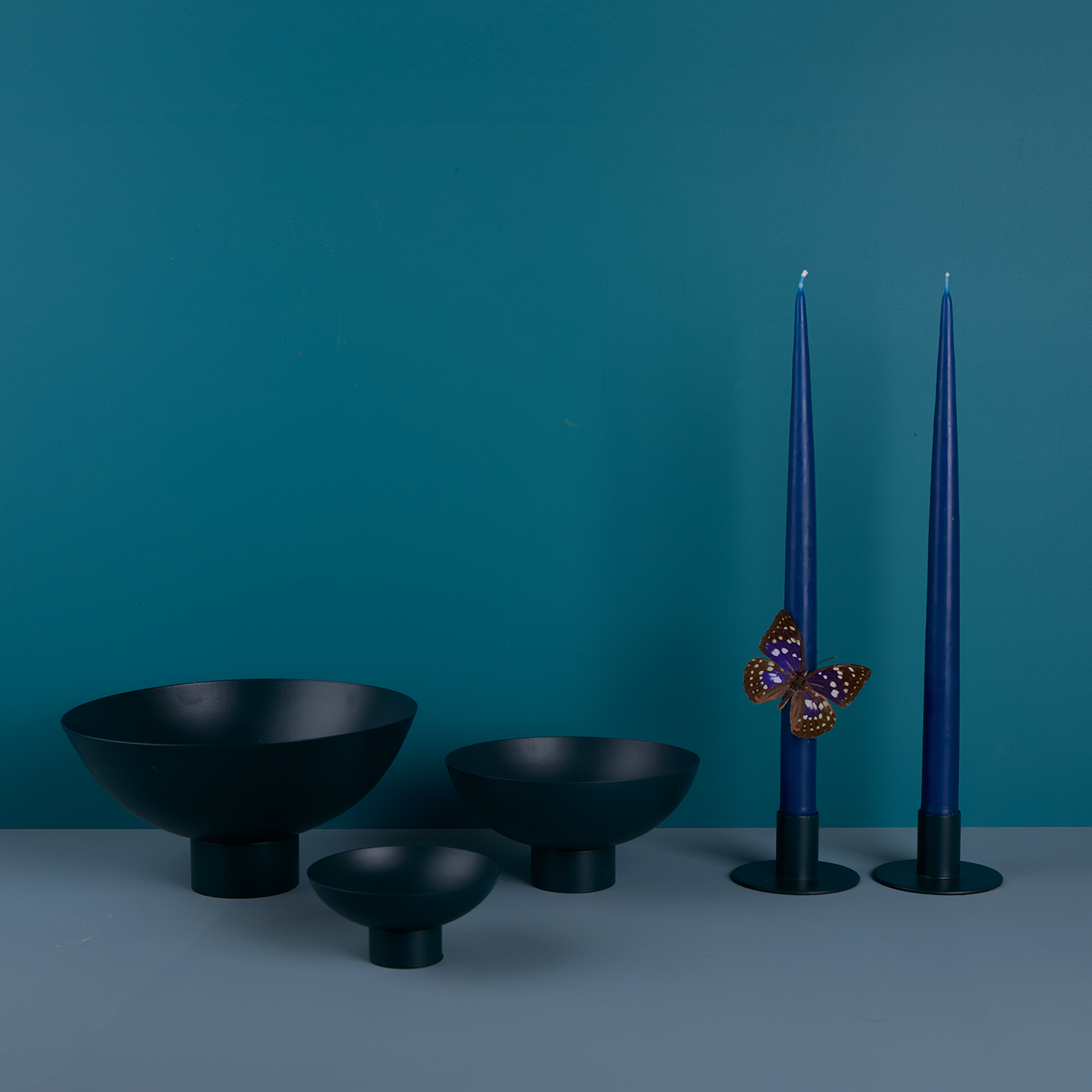 A trio of footed bowls and pair of candle holders, all in Peacock, with blue taper candles. Styled with a butterfly.