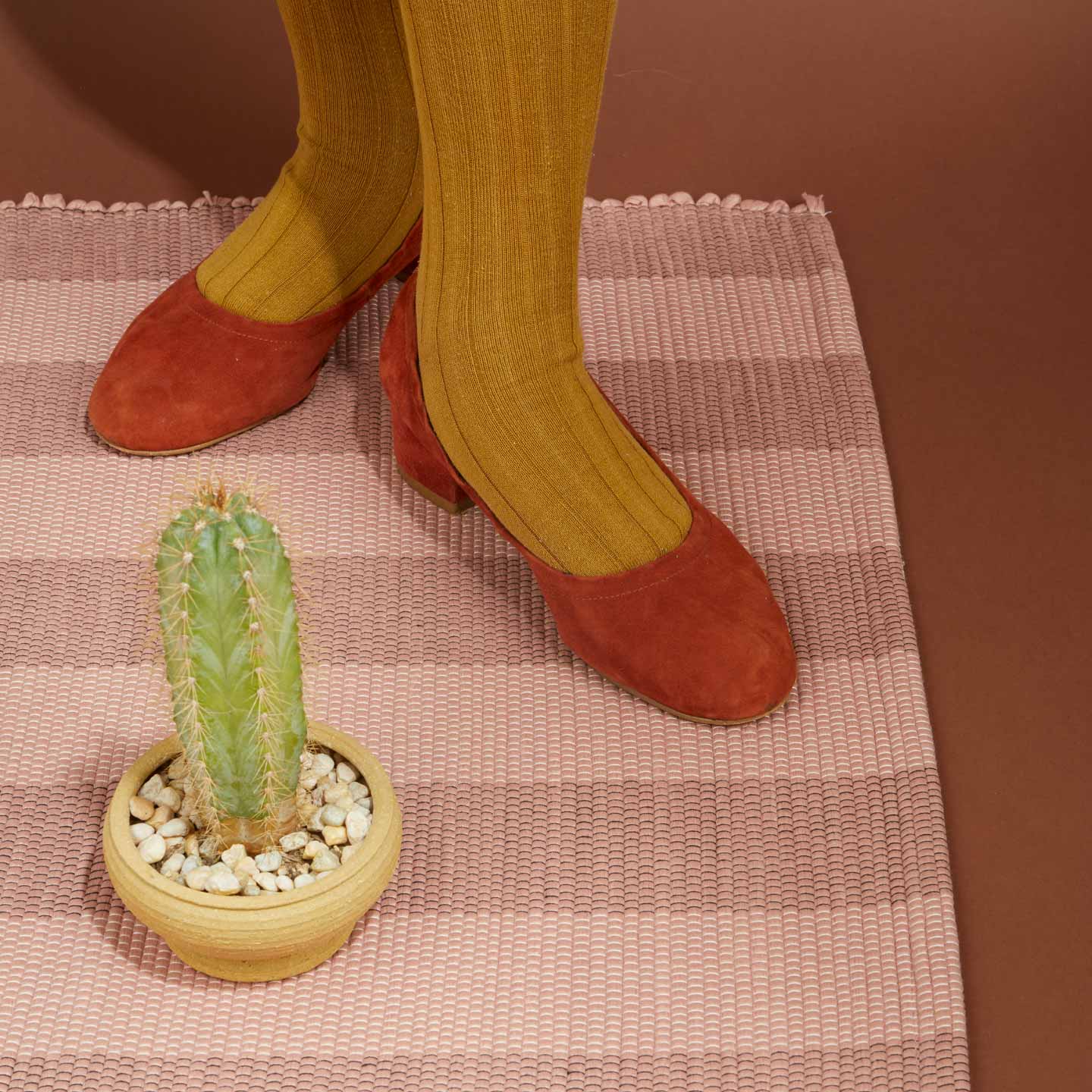 Person wearing mustard yellow socks and red shoes standing on Essential Floor Mat in Blush with a small potted cactus.