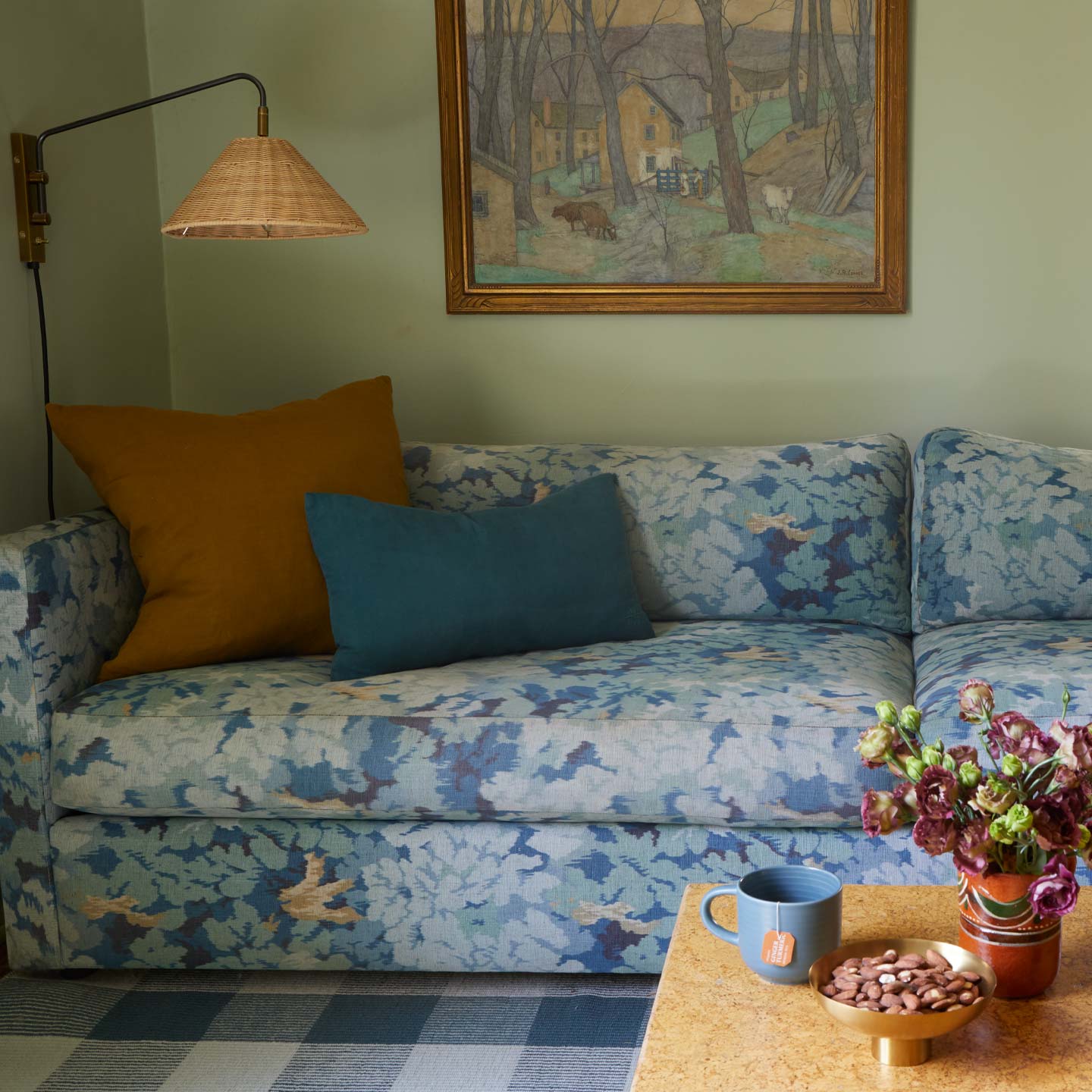 Den with floral sofa, art, wall lamp, and coffee table with mug, nuts, and vase.
