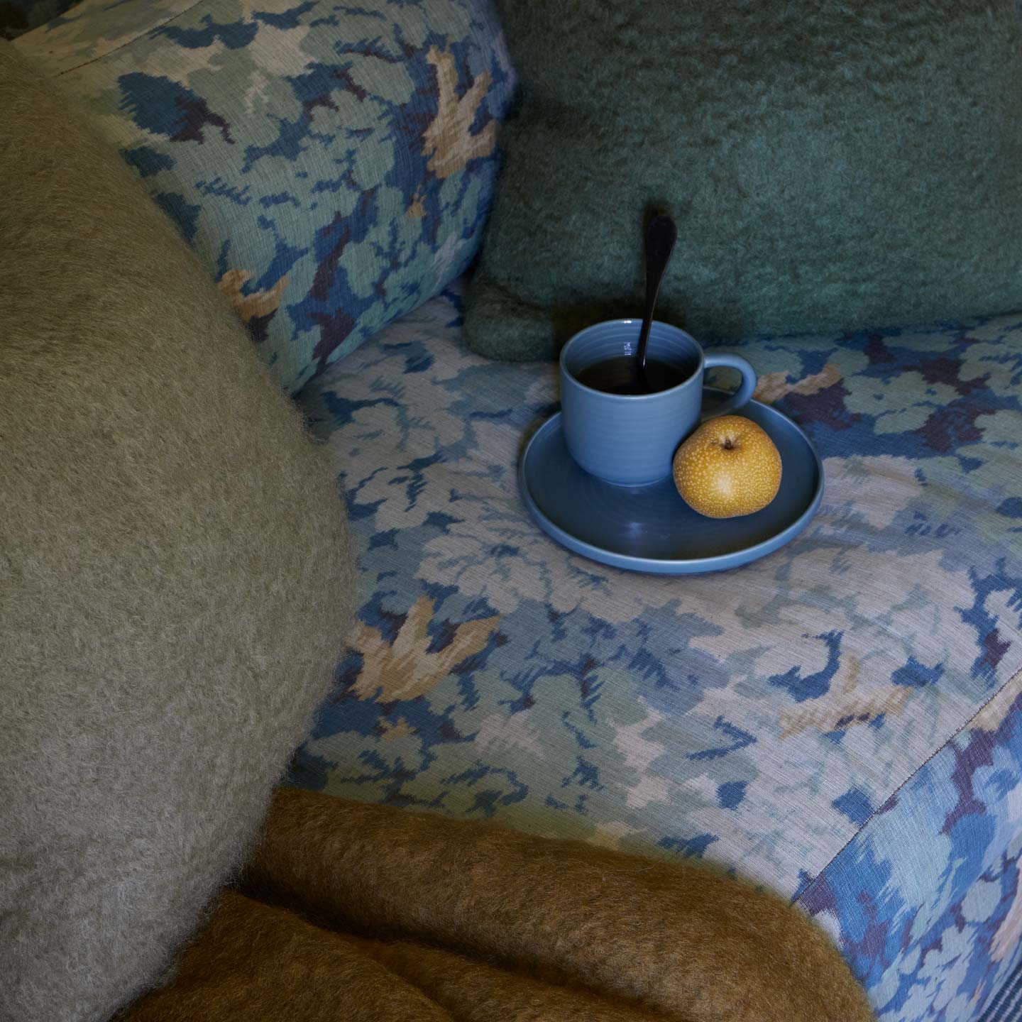 Blue mug with a spoon and an apple on a patterned surface next to a textured wall.
