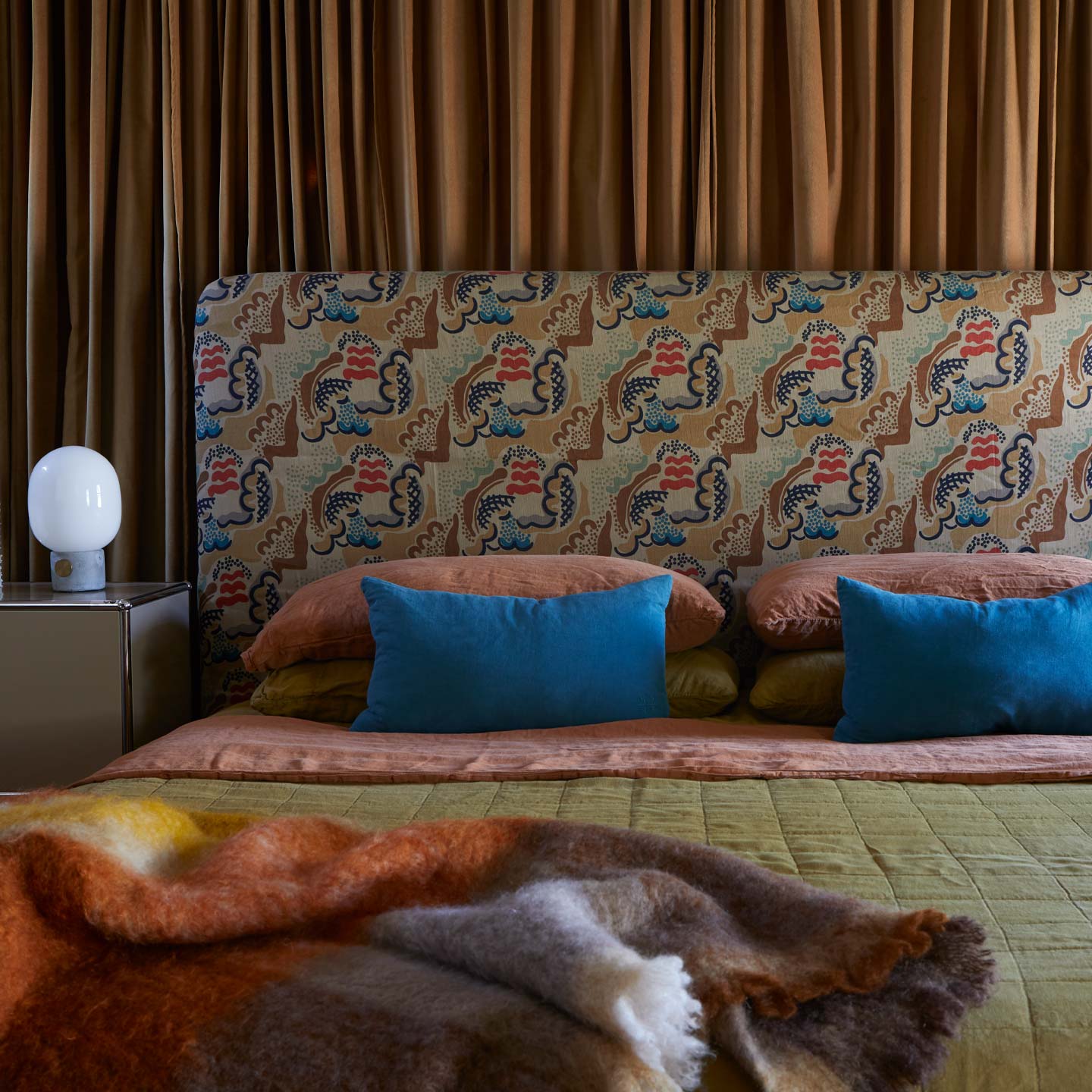 Bed with patterned headboard, terracotta, bronze, and peacock bedding with multicolor throw.