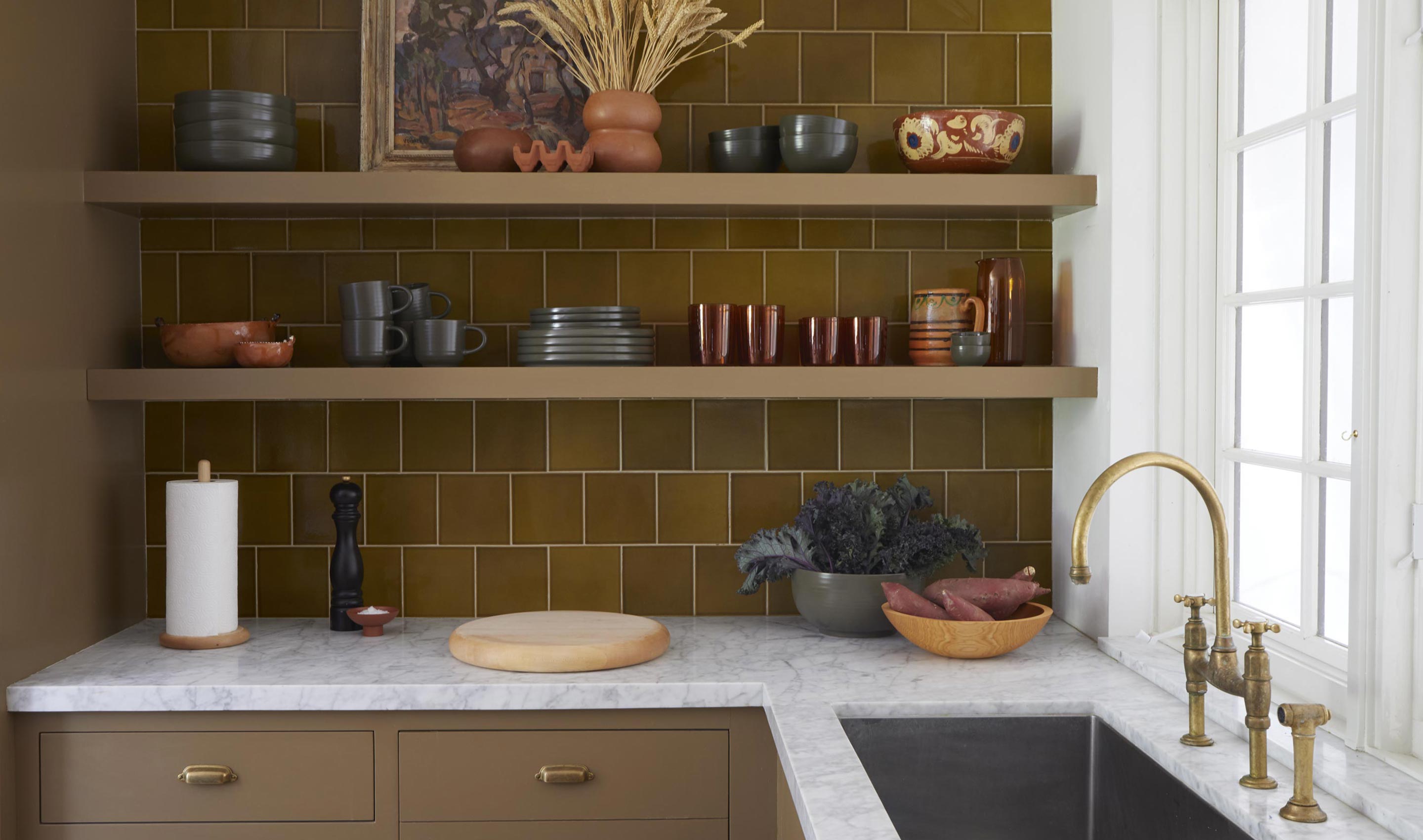 A kitchen scene with shelves against a green tiled wall stacked with dinnerware, glassware, bowls, vases and pitchers. The white marble countertop is arranged with bowls of produce, a chopping block, and kitchen accessories.