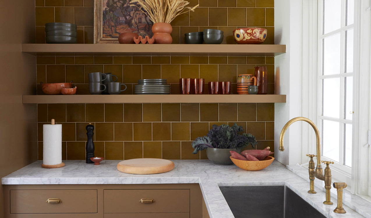 A kitchen scene with shelves against a green tiled wall stacked with dinnerware, glassware, bowls, vases and pitchers. The white marble countertop is arranged with bowls of produce, a chopping block, and kitchen accessories.