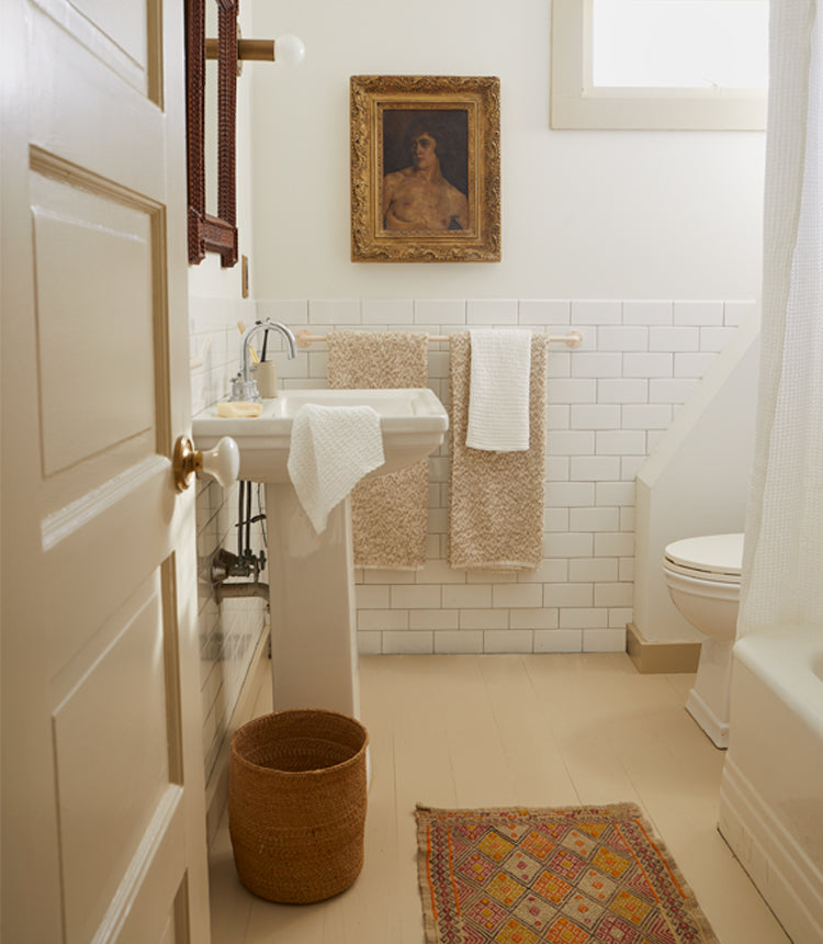 A small bathroom with white tiled walls, a pedestal sink, a toilet, a wicker basket, two towels on a rack, and a framed painting above the sink. A small rug is placed on the floor.