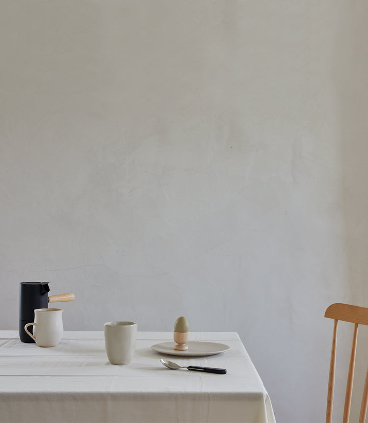 A minimalist table setting with a black kettle, a white pitcher, a mug, a plate with an egg in an egg cup, and a knife. A wooden chair is partially visible on the right side.