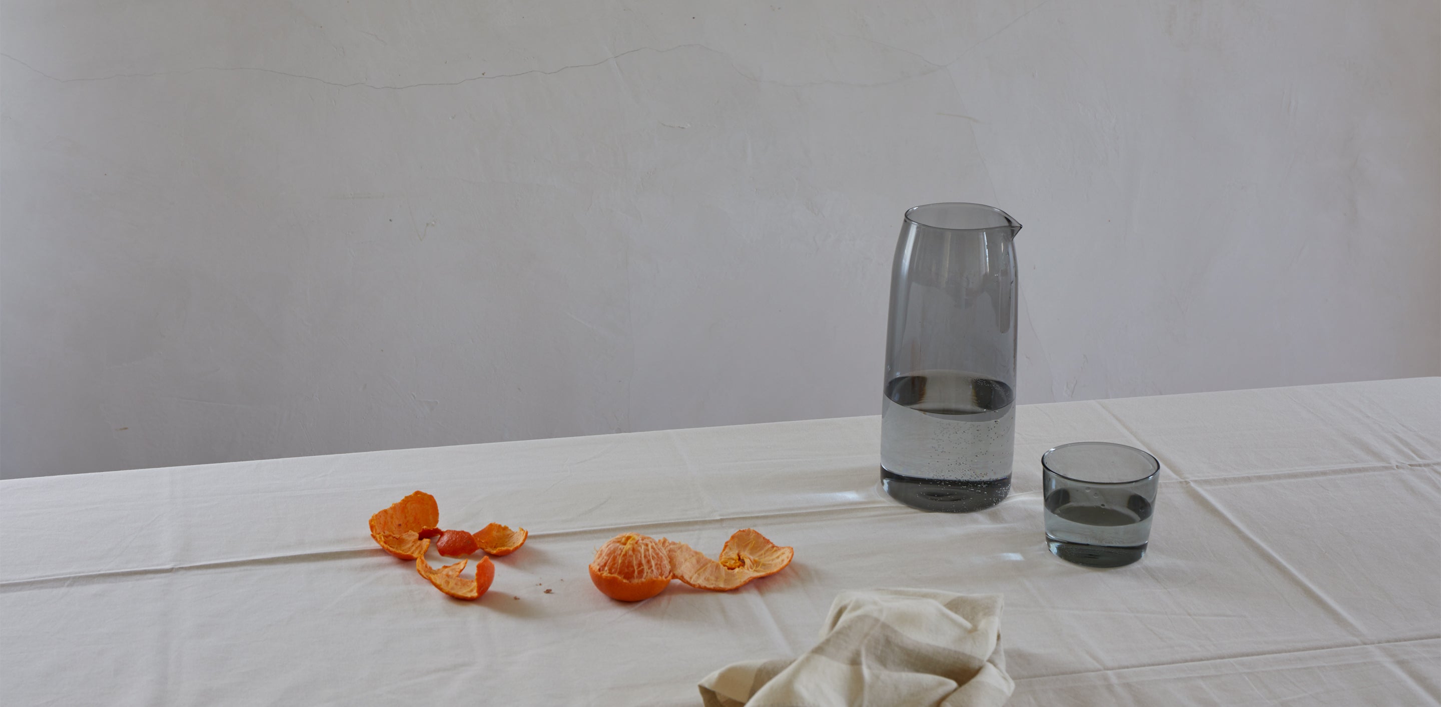 A table with a white cloth holds an empty tangerine peel, a beige napkin, a half-filled glass carafe, and a glass of water.