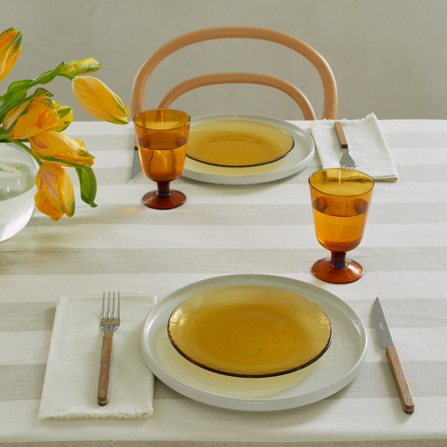 Place setting with amber and ivory plates, amber glassware with water, flowers, and teak flatware on ivory napkins.
