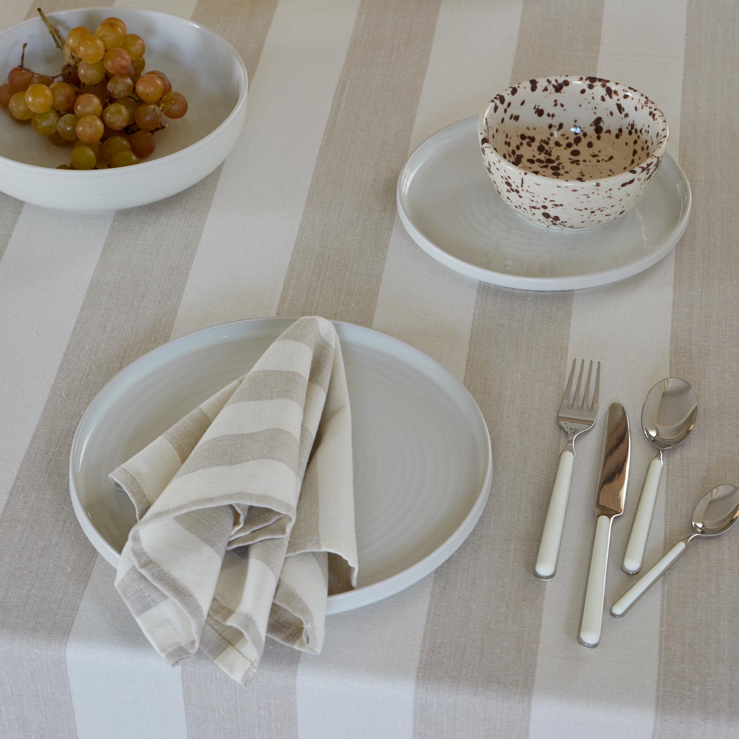 Place setting with Essential Dinnerware and Essential Striped Napkin in Ivory and Essential Striped Tablecloth in Ivory with flatware and grapes.