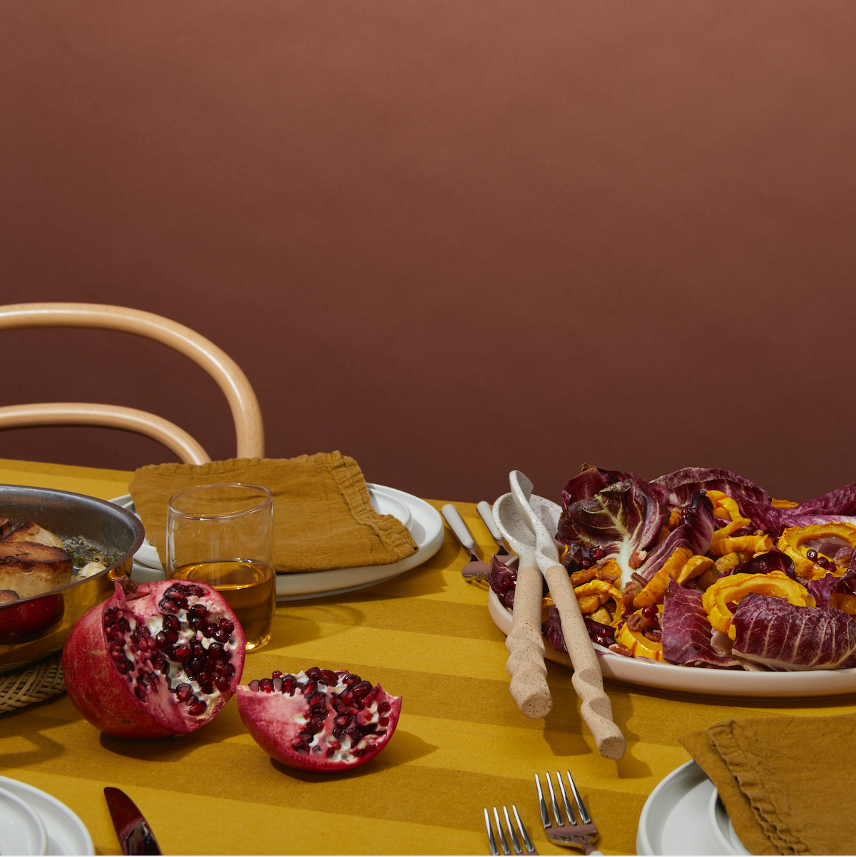 A table set with plates, cutlery, a glass, a pomegranate, and a bowl of sliced vegetables. The background wall is brown.