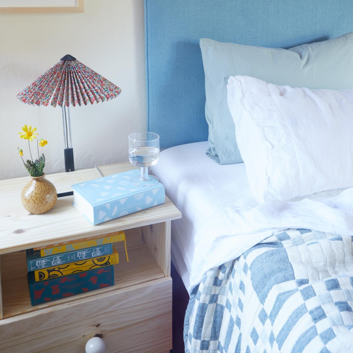 Bedroom with a nightstand, lamp, and books near a bed with white bedding and blue headboard.