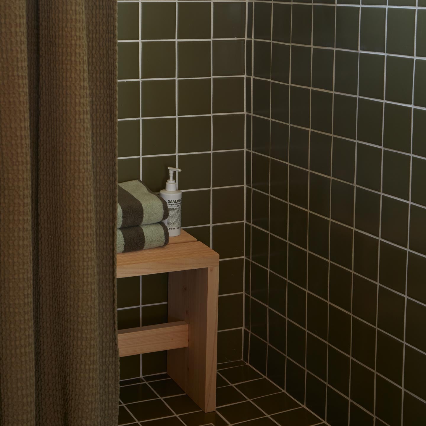 Bathroom corner with dark tiled walls, a wooden shelf with towels and a bottle, and a shower curtain.