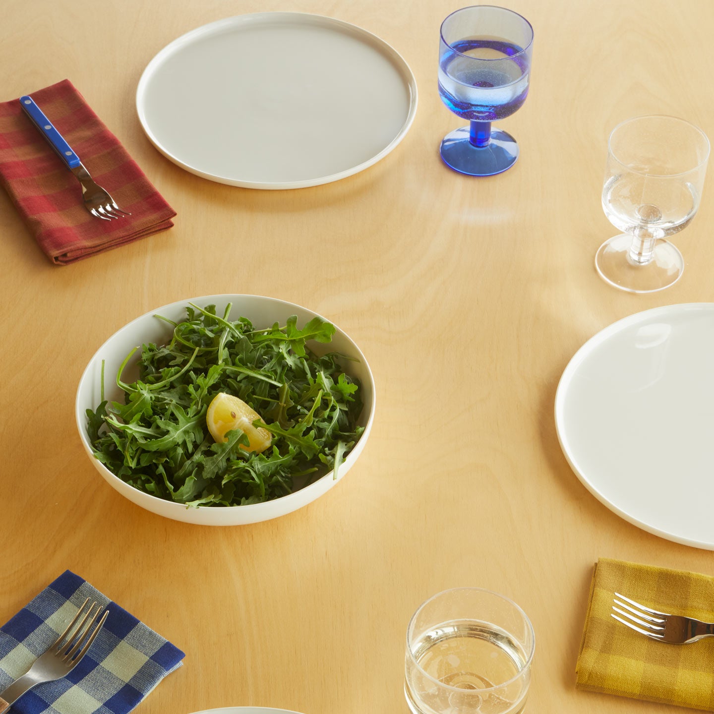 Table setting with Simple Porcelain Plates, salad in Simple Porcelain Low Bowl, Essential Gingham Napkins in Mustard Red, and Blue, forks, and Simple Seeded Glassware in Clear and Blue.