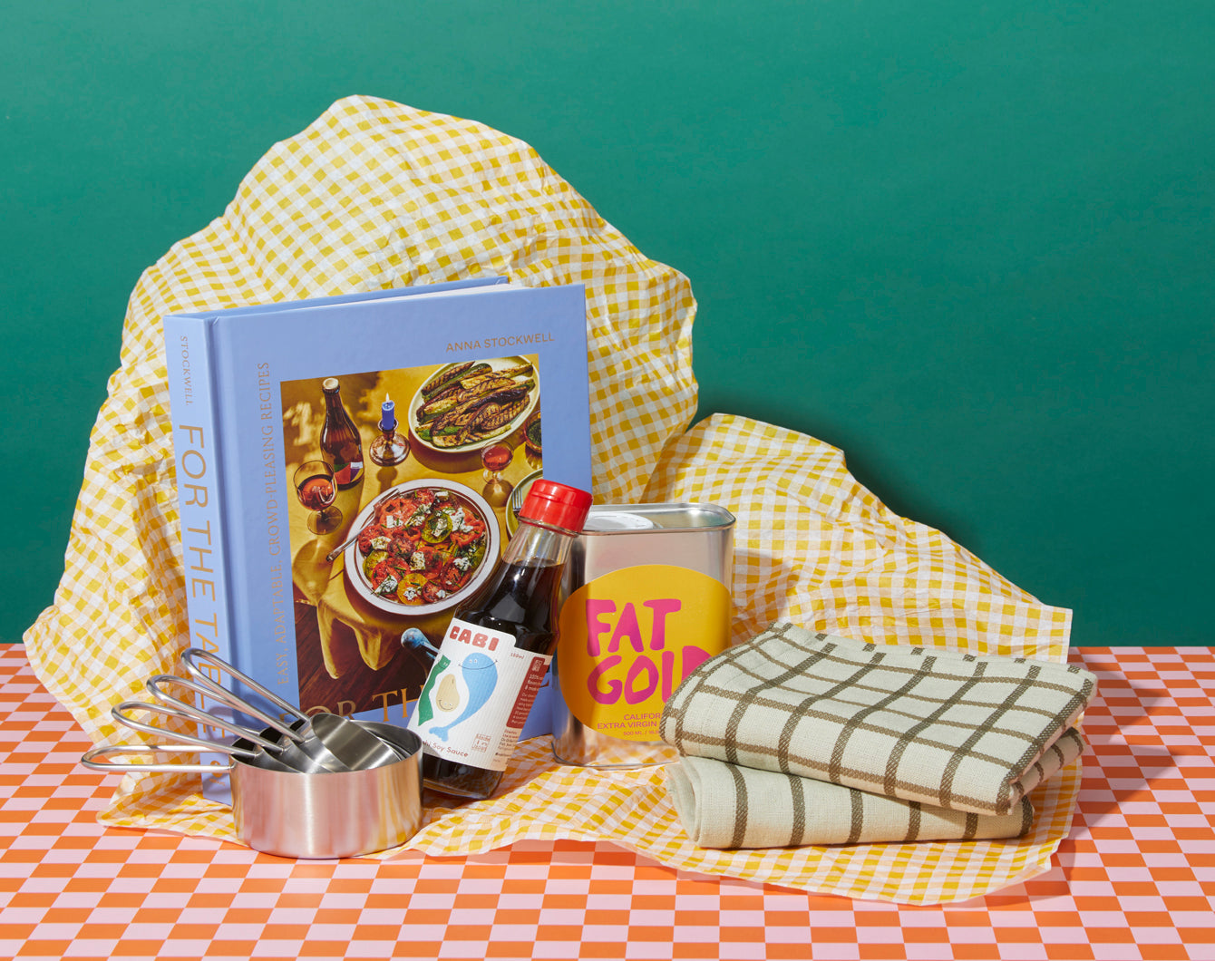A cookbook, measuring cups, a bottle of soy sauce, a tin labeled Fat Gold, and dish towels are arranged on a checkered table with a yellow checkered fabric backdrop against a green background.