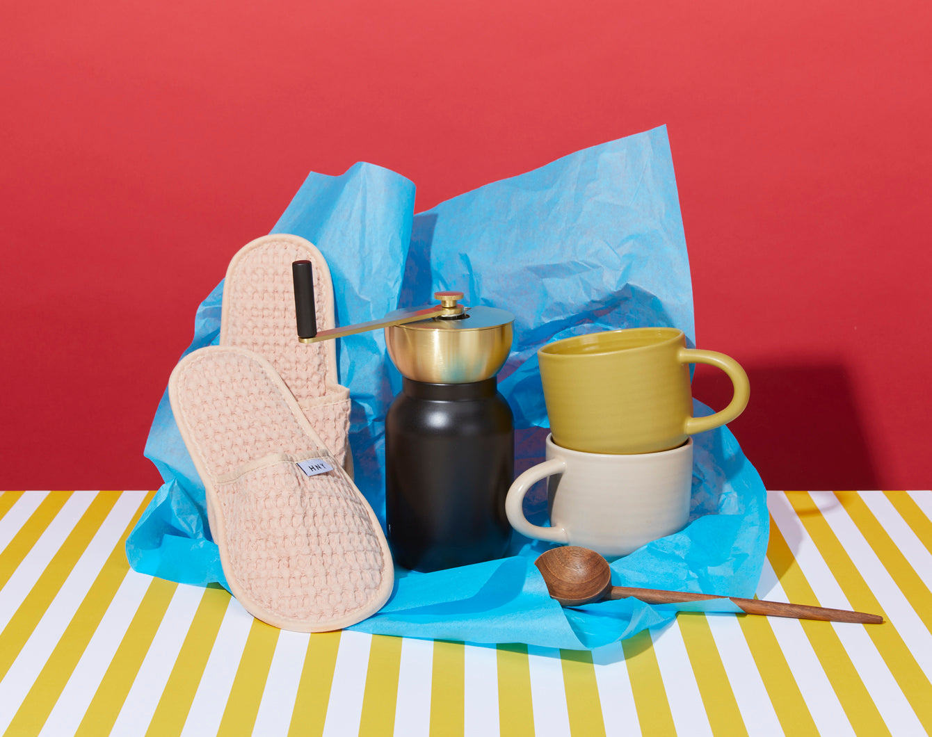 A pair of slippers, a coffee grinder, a yellow and a white mug, and a honey dipper are arranged on blue tissue paper against a red background with a yellow and white striped surface.