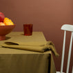 Table setting with olive tablecloth and napkin and footed bowl with fruit and glass with water and white chair with brown background.