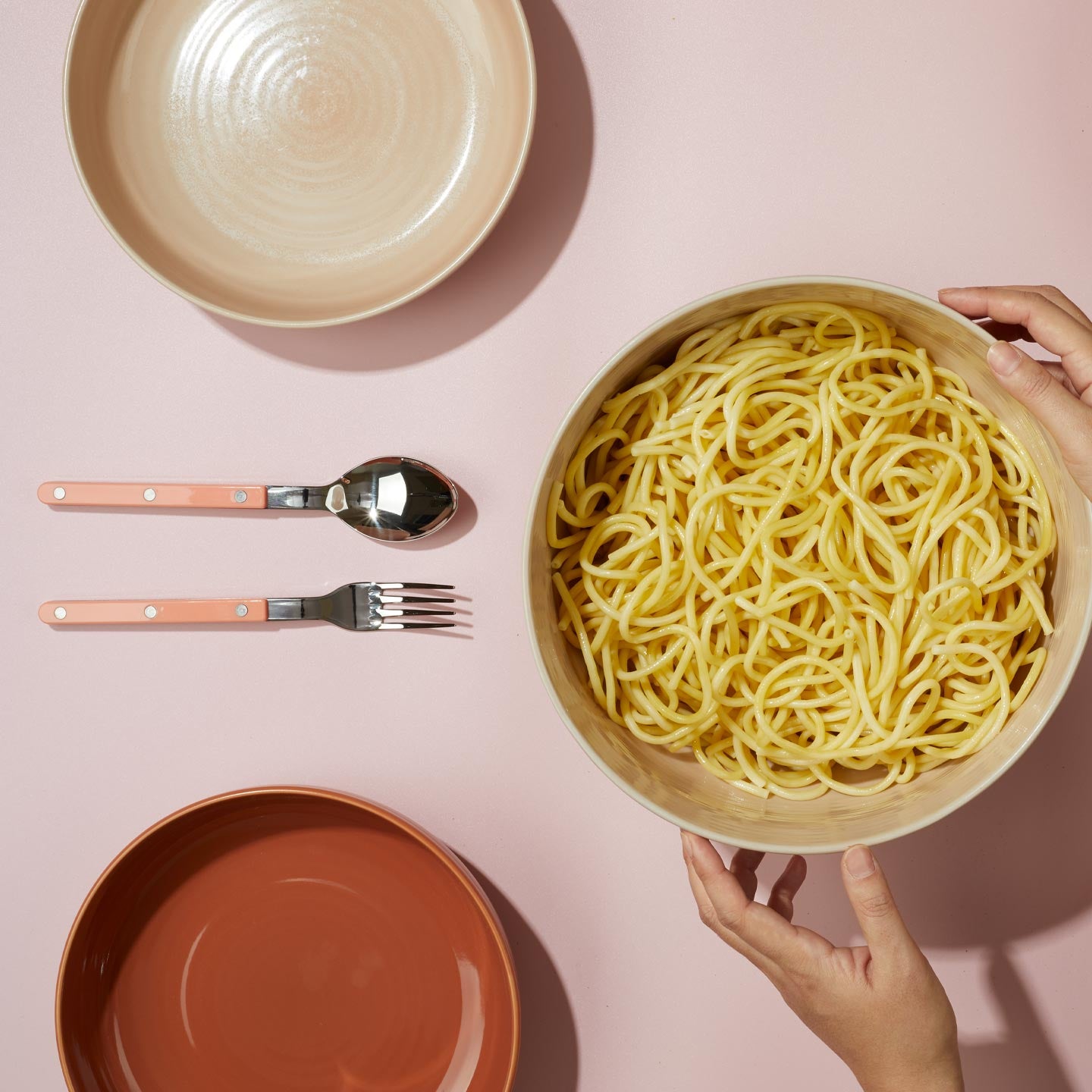 Essential serving bowl of spaghetti held by a hand on a pink background with other ceramic bowls and cutlery.
