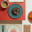 Chocolate donut with sprinkles on a peacock essential plate next to a cup of coffee on a red surface.