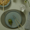 Green artichoke on a blue plate with chopsticks, set on a light green tablecloth.