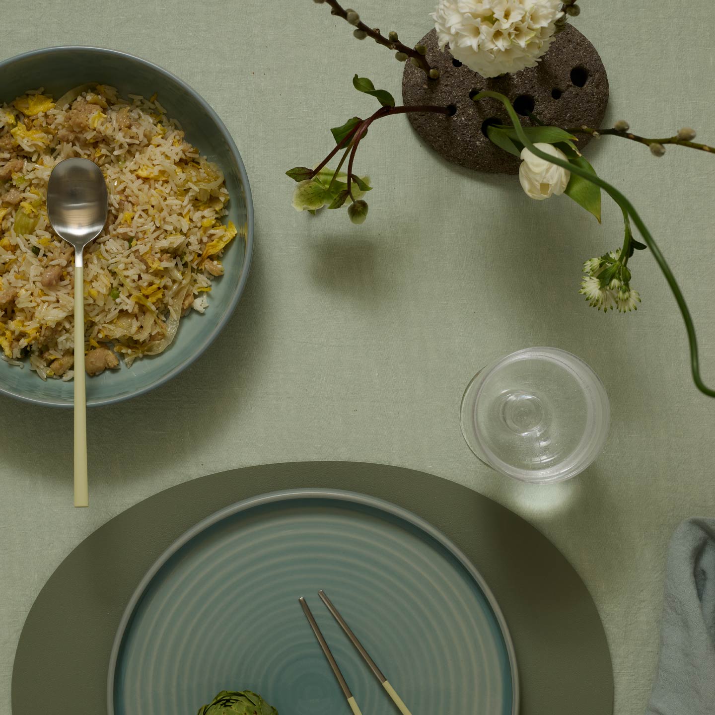Table setting with a bowl of food, a plate, chopsticks, and a glass on a green tablecloth.