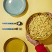 Person holding an essential serving bowl of spaghetti with a yellow background, including a blue plate and silverware.