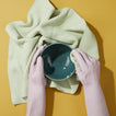 Hands in pink gloves washing Peacock Cereal bowl with Sage Waffle Dishcloth on yellow background.