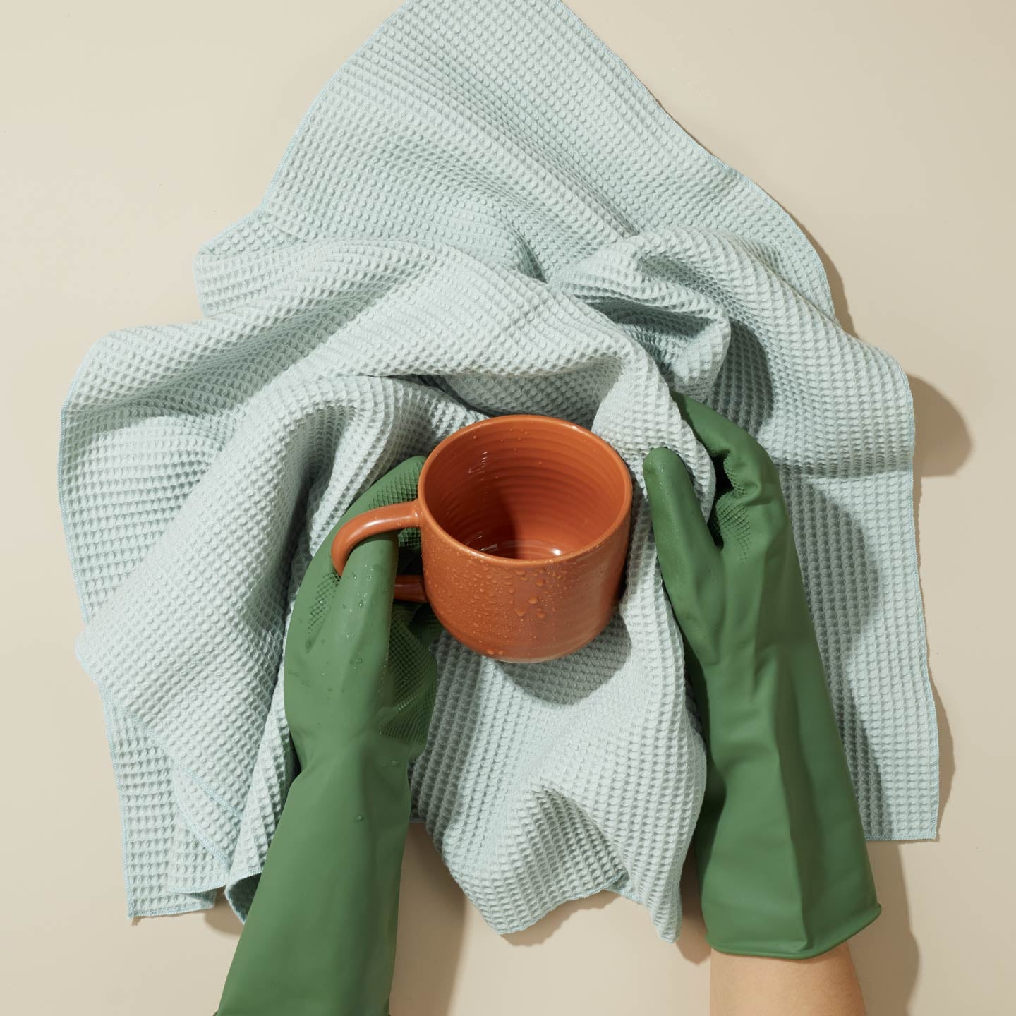 Person holding a terracotta cup with a green glove, on a sky Essential towel on a beige background