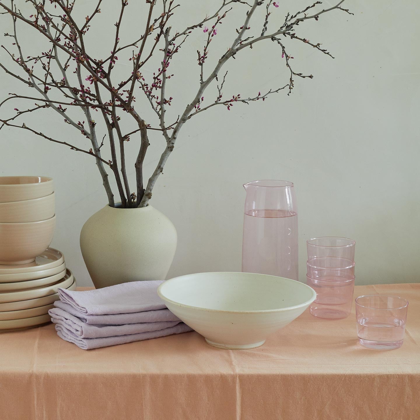 Table with stack of Essential Dinnerware in blush, vase with branches, Lilac napkins, and Essential Glass Pitcher and Glassware in Blush.