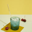 Peacock essential glass with ice and a straw on a yellow cocktail napkin, with cherries on a light background