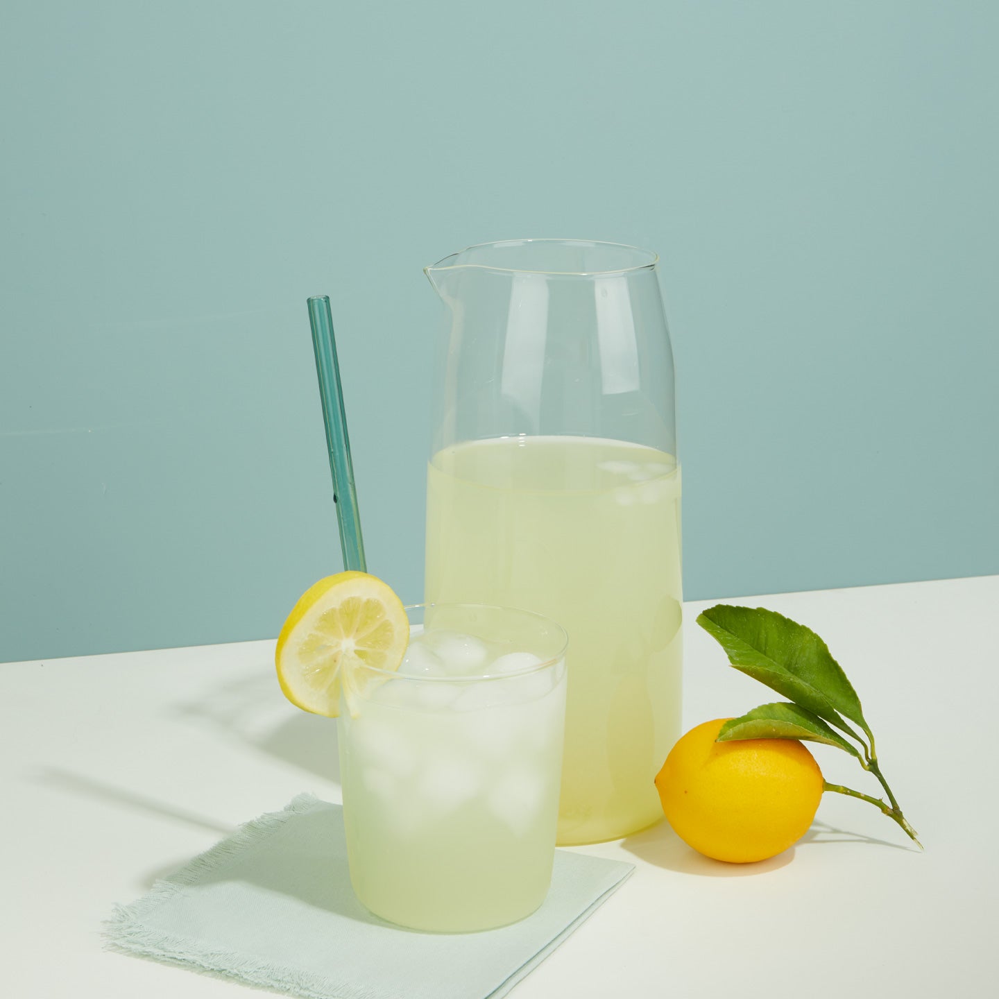 Glass of lemonade with ice and a pitcher of lemonade on a light blue background