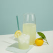 Glass of lemonade with ice and a pitcher of lemonade on a light blue background