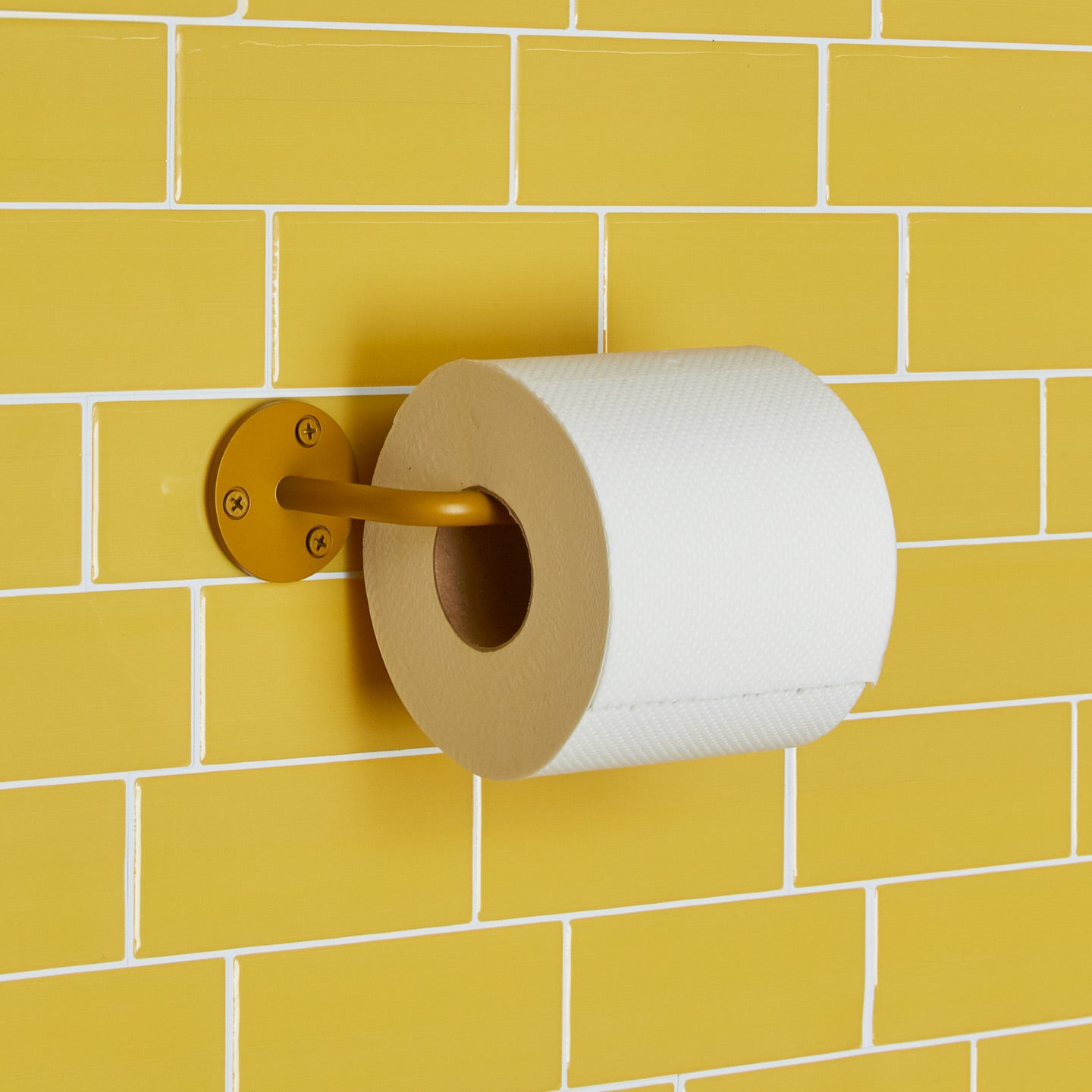 Essential toilet paper holder in Mustard with a roll of toilet paper on tiled wall.