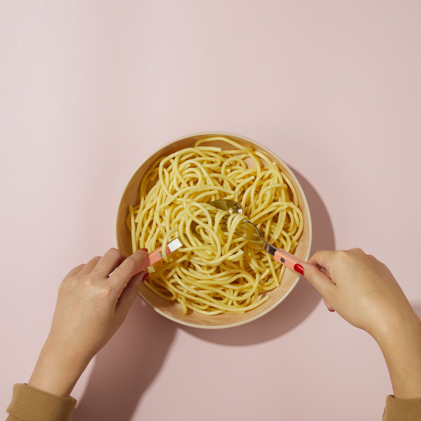 Hands scooping pasta with flatware from bowl on pink background.