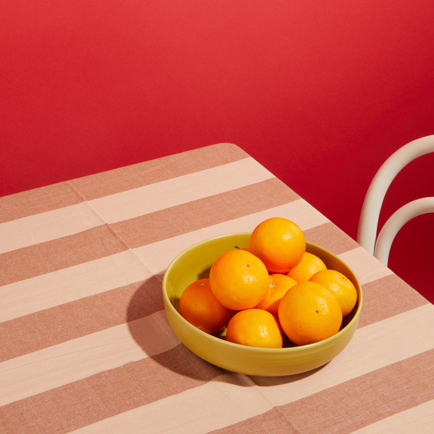 Oranges in Mustard Essential Low Bowl on Striped blush Tablecloth with chair and red background.