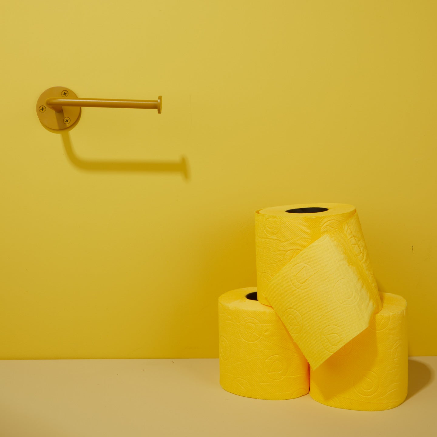 Three rolls of yellow toilet paper on a beige surface with a yellow wall background and mustard toilet paper holder.