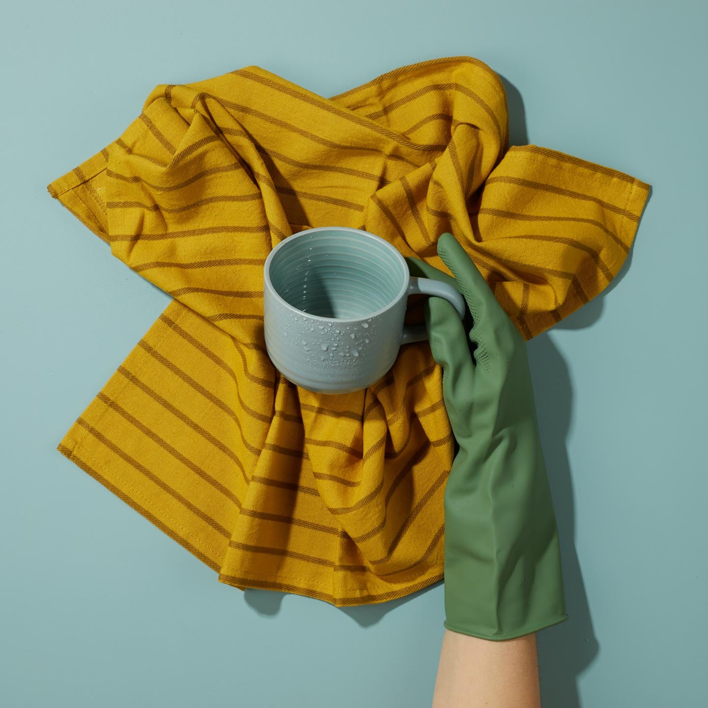 Hand wearing green glove holding wet blue mug over Essential Mustard Dishtowel on blue background.