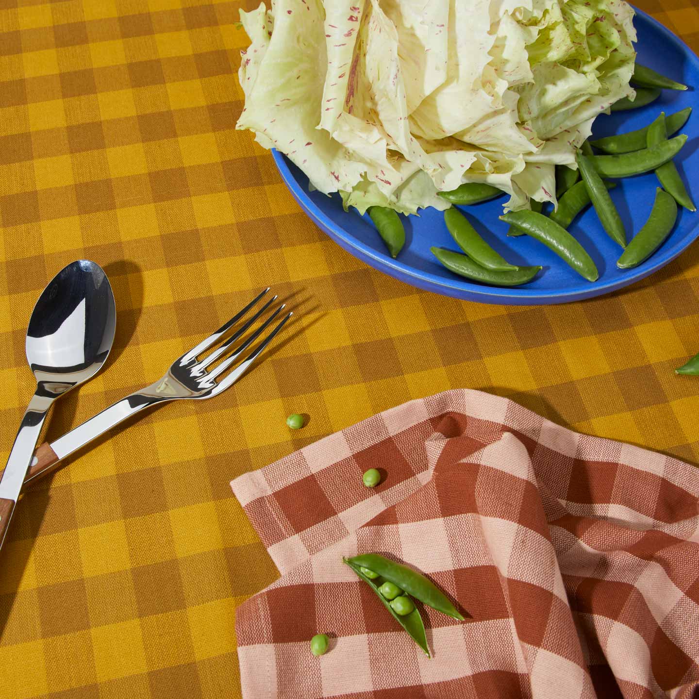Essential Gingham napkin in Blush on Essential Gingham tablecloth in Mustard with Serving Set and blue plate with peas and greens.