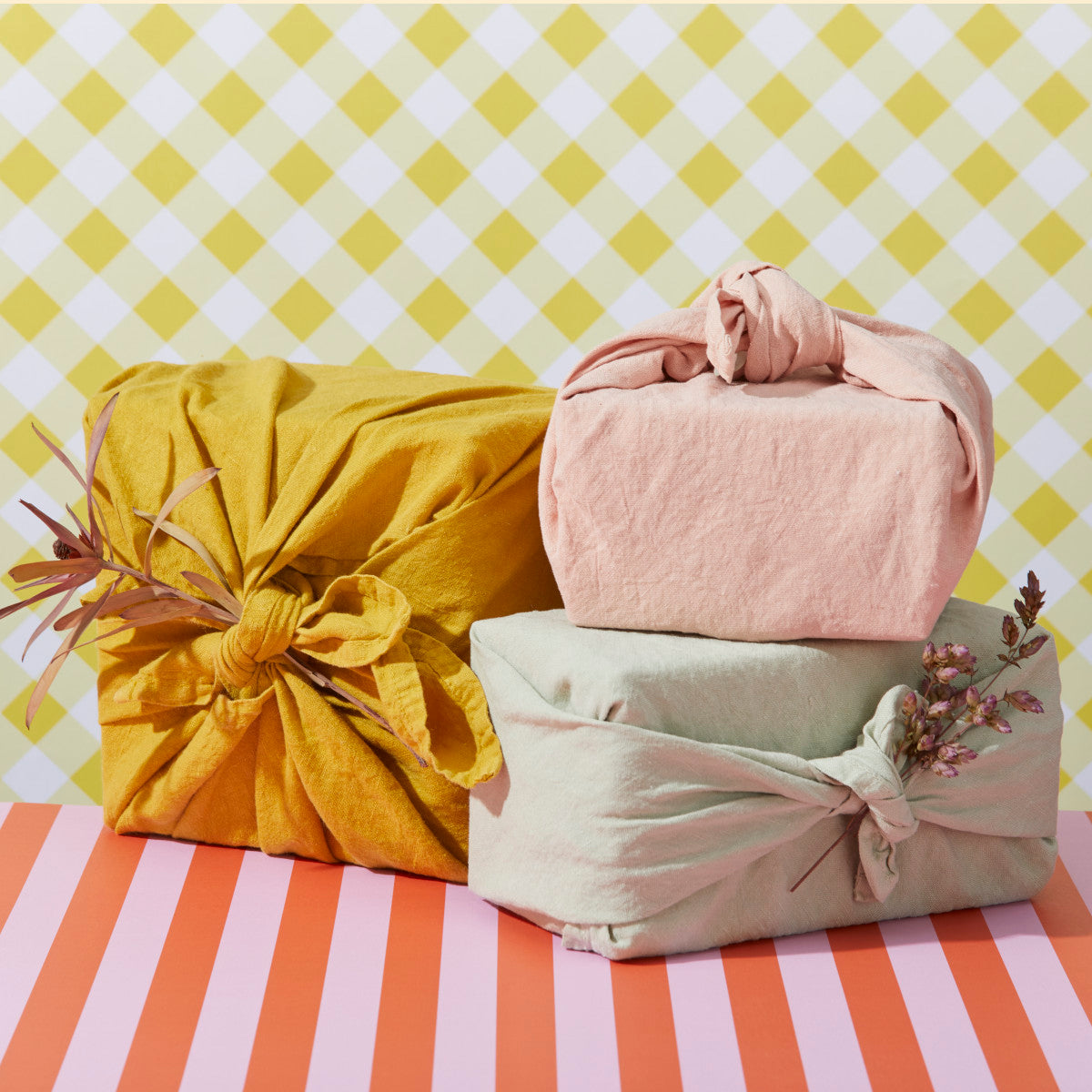 Three fabric-wrapped gifts, one in yellow, one in pink, and one in light green, with decorative plants attached, are displayed against a checkered yellow background and a striped pink surface.