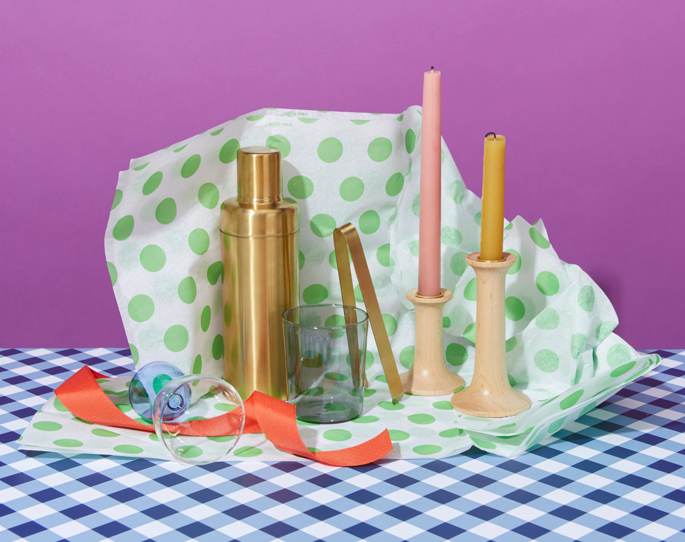 A gold cocktail shaker, clear glass, pink and yellow candles on holders, ribbon, and sunglasses are arranged on a green polka dot paper over a table with blue checkered cloth, with a purple background.