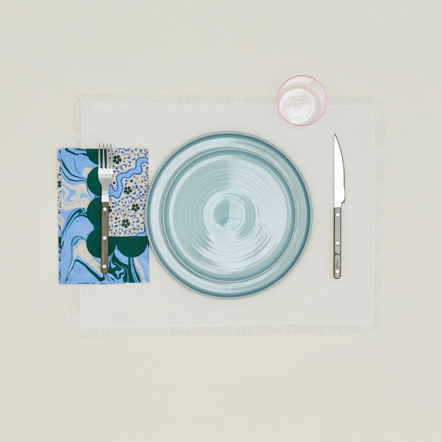 A table setting with sky plates, a blush glass, an ivory placemat, and a cornflower patterned napkin.