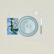 A table setting with sky plates, a blush glass, an ivory placemat, and a cornflower patterned napkin.