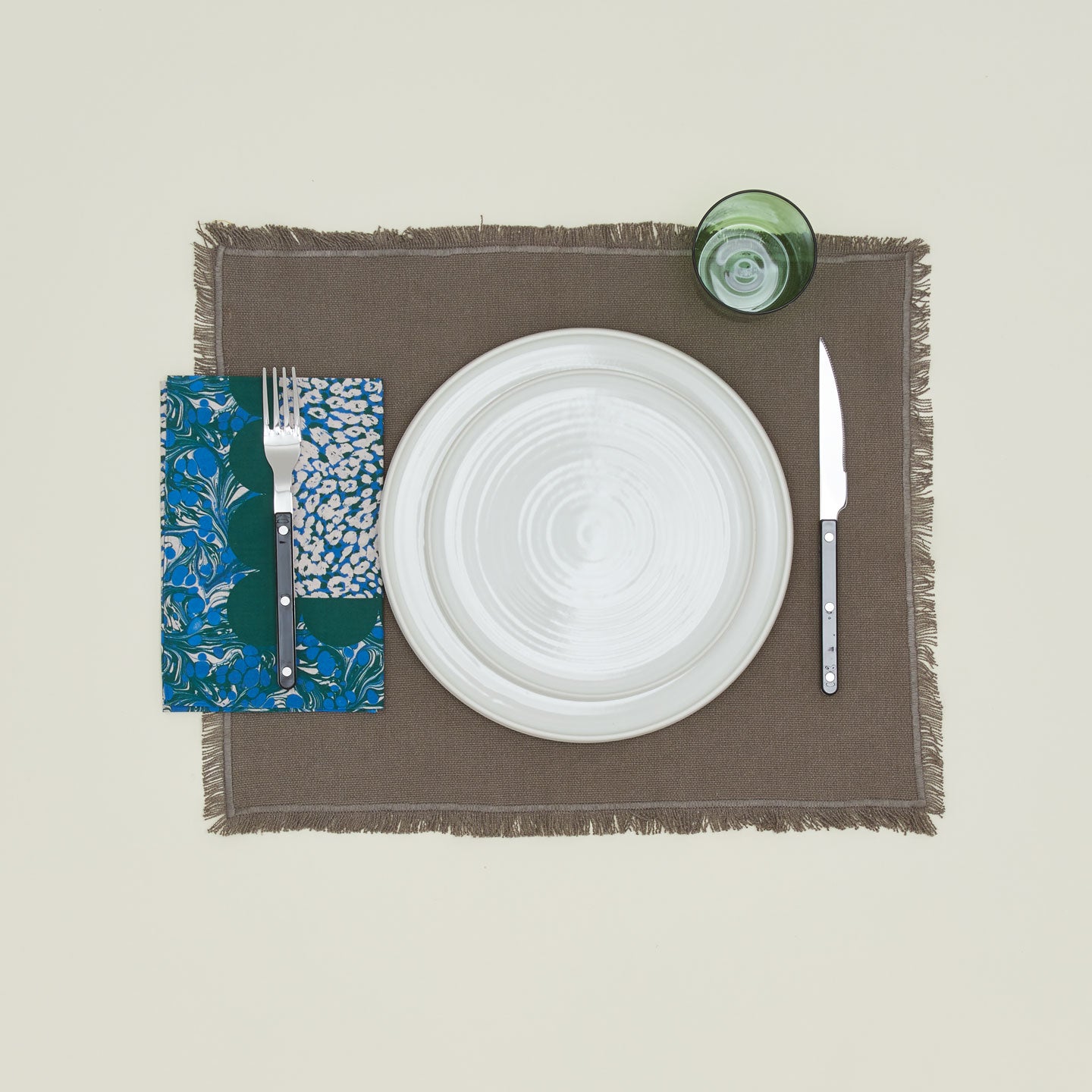 A table setting with bone plates, a green glass, a grey placemat, and an evergreen patterned napkin.